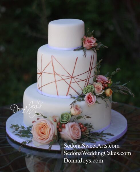 Three Tier Hand Painted rose gold Geometric lines painted on center tier. Adorned with combo of fresh flowers,Sugar Succulents & Rose Gold Sugar Berries. Created by Pastry Chef Donna Joy.